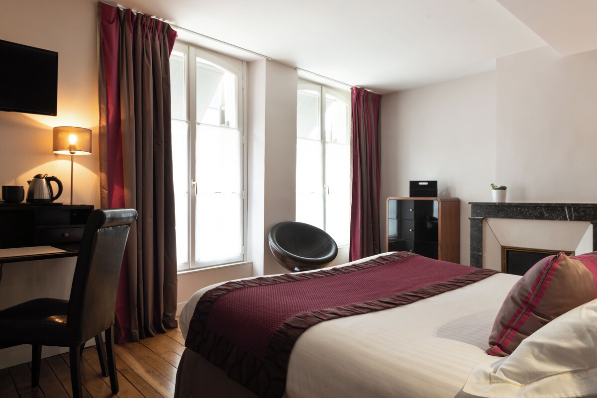 A hotel room with a bed, desk, chair, TV, and a fireplace.