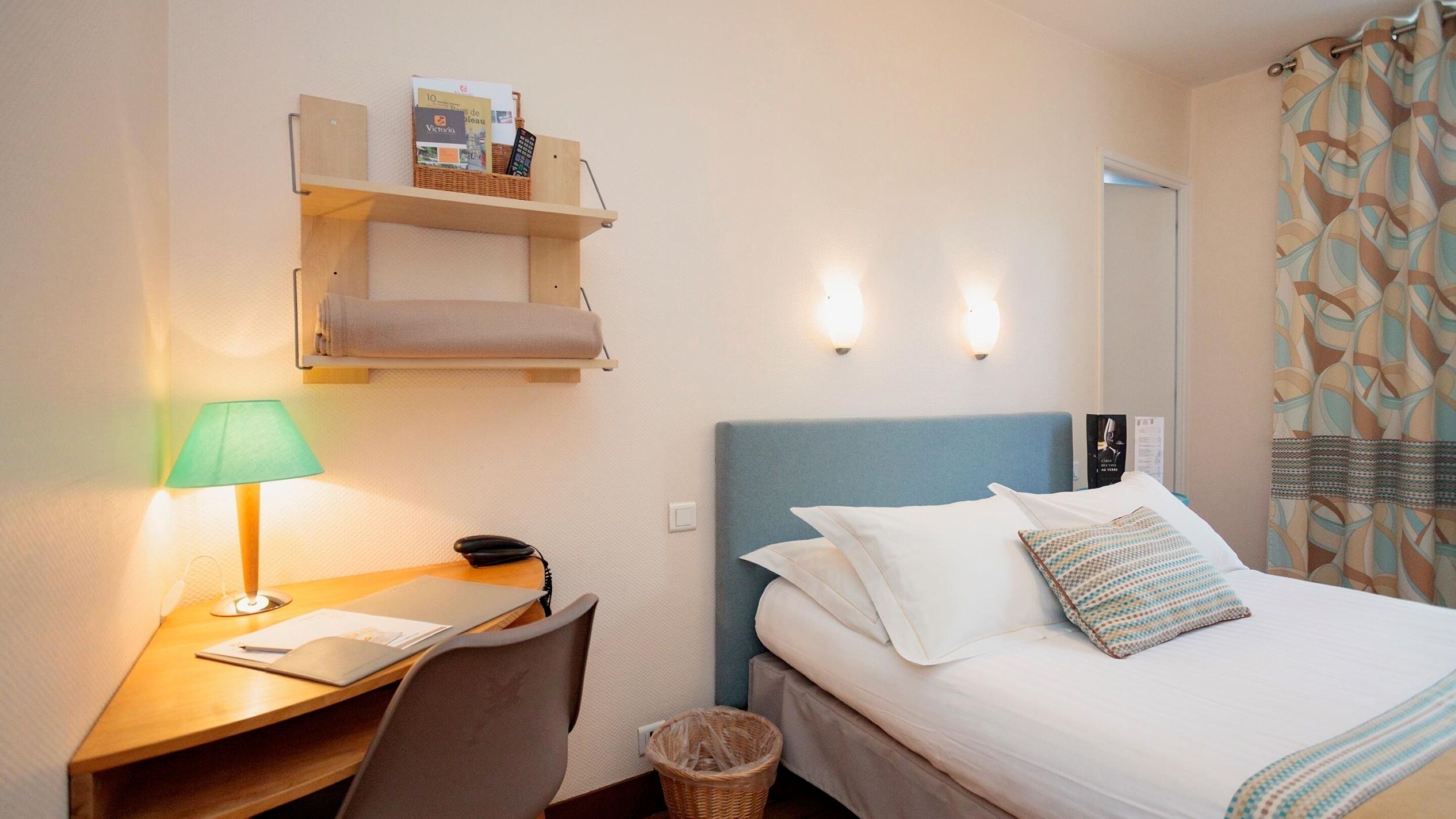 A hotel room with a bed, a desk with a lamp, a chair, a small shelf with books and a remote control, and a trash bin.