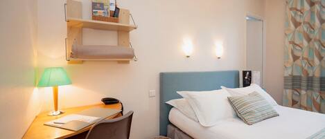 A hotel room with a bed, a desk with a lamp, a chair, a small shelf with books and a remote control, and a trash bin.