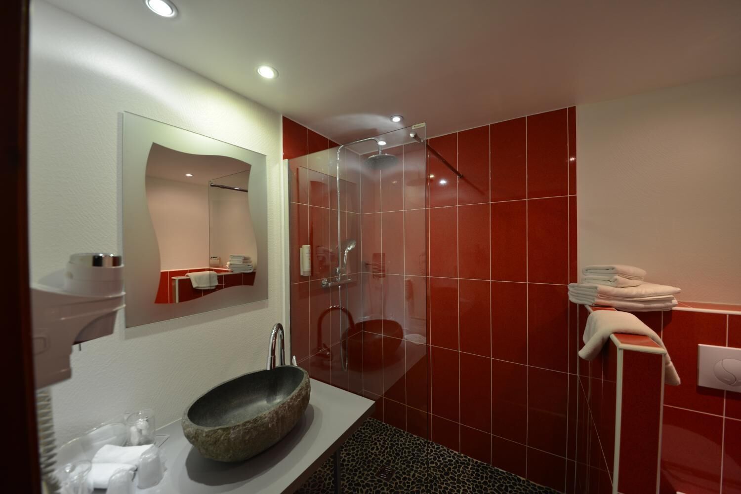 A modern bathroom with a large mirror, a stone basin, a glass shower enclosure, and red tiled walls.