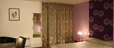A hotel room with a bed, a desk, a chair, a nightstand with a lamp, and a wall with a circular pattern.