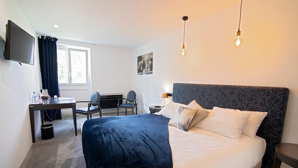 A hotel room with a large bed, a desk, two chairs, a flat-screen TV, and a window with curtains.