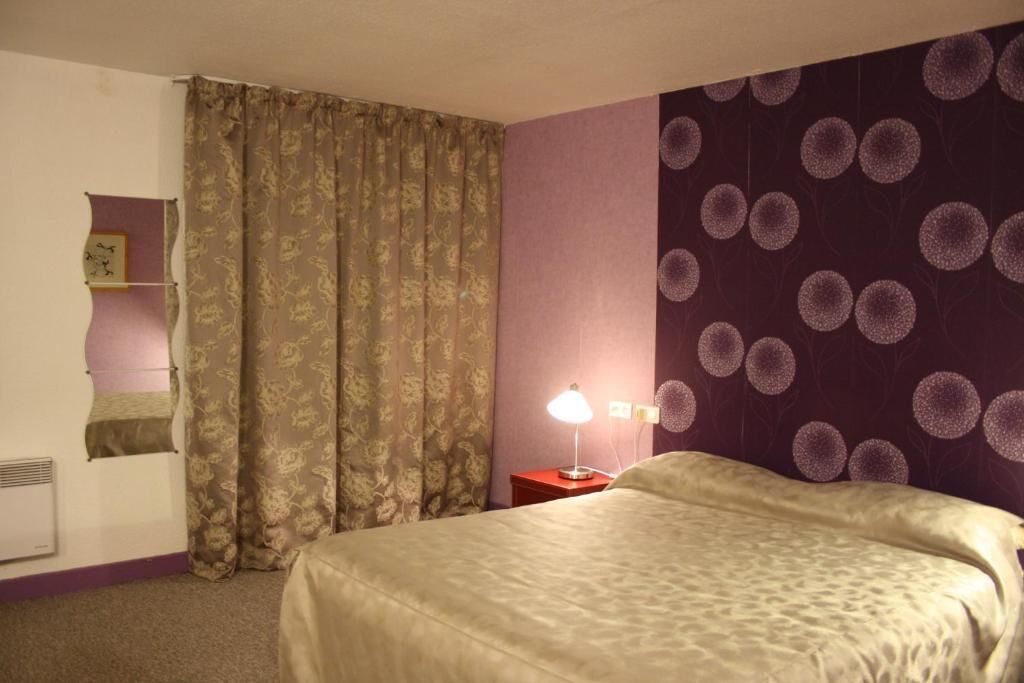 Une chambre d’hôtel avec un lit, une table de chevet avec une lampe, une étagère fixée au mur et une fenêtre avec des rideaux.