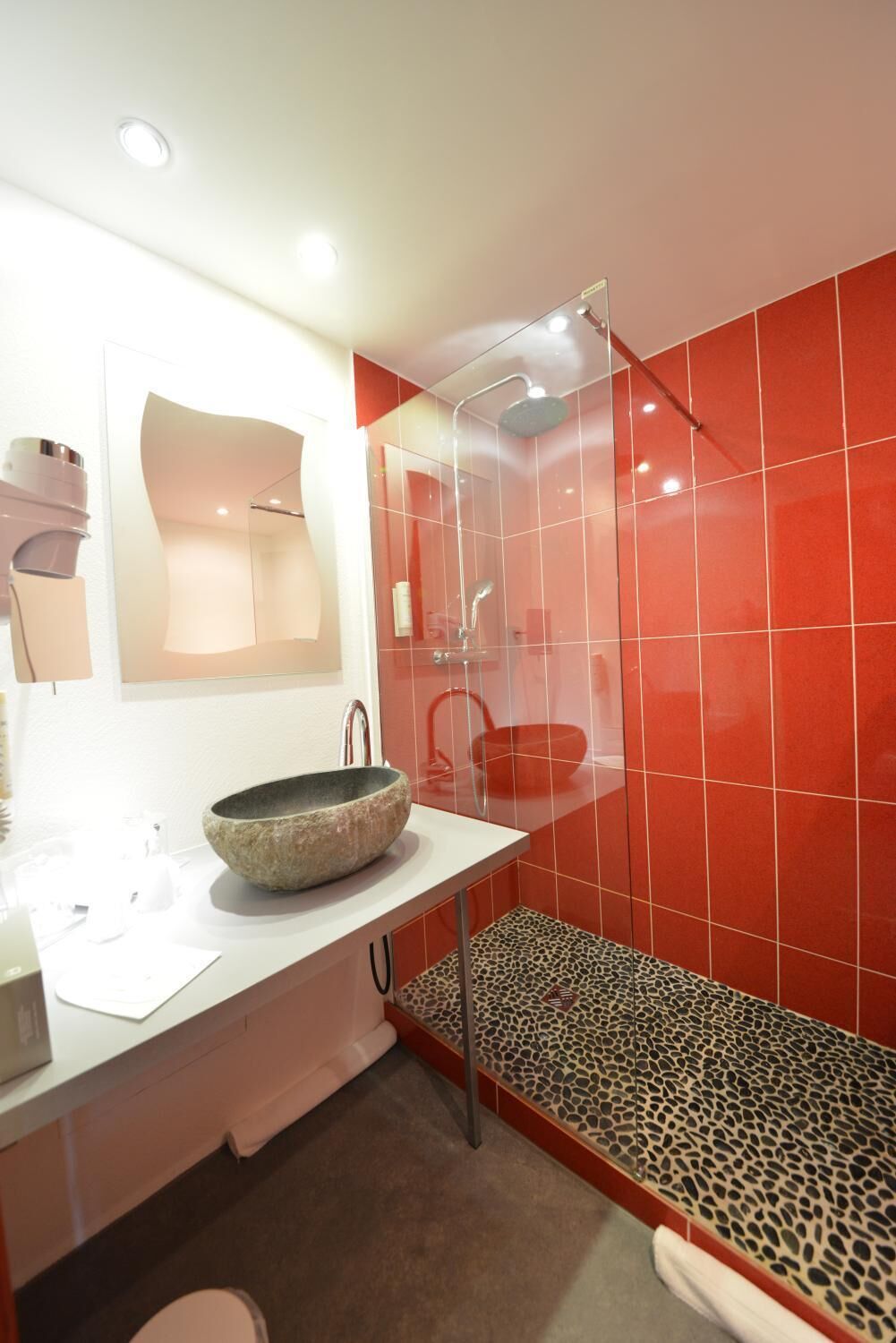 A modern bathroom with a white sink, a stone basin, a glass shower enclosure, and red tiled walls.
