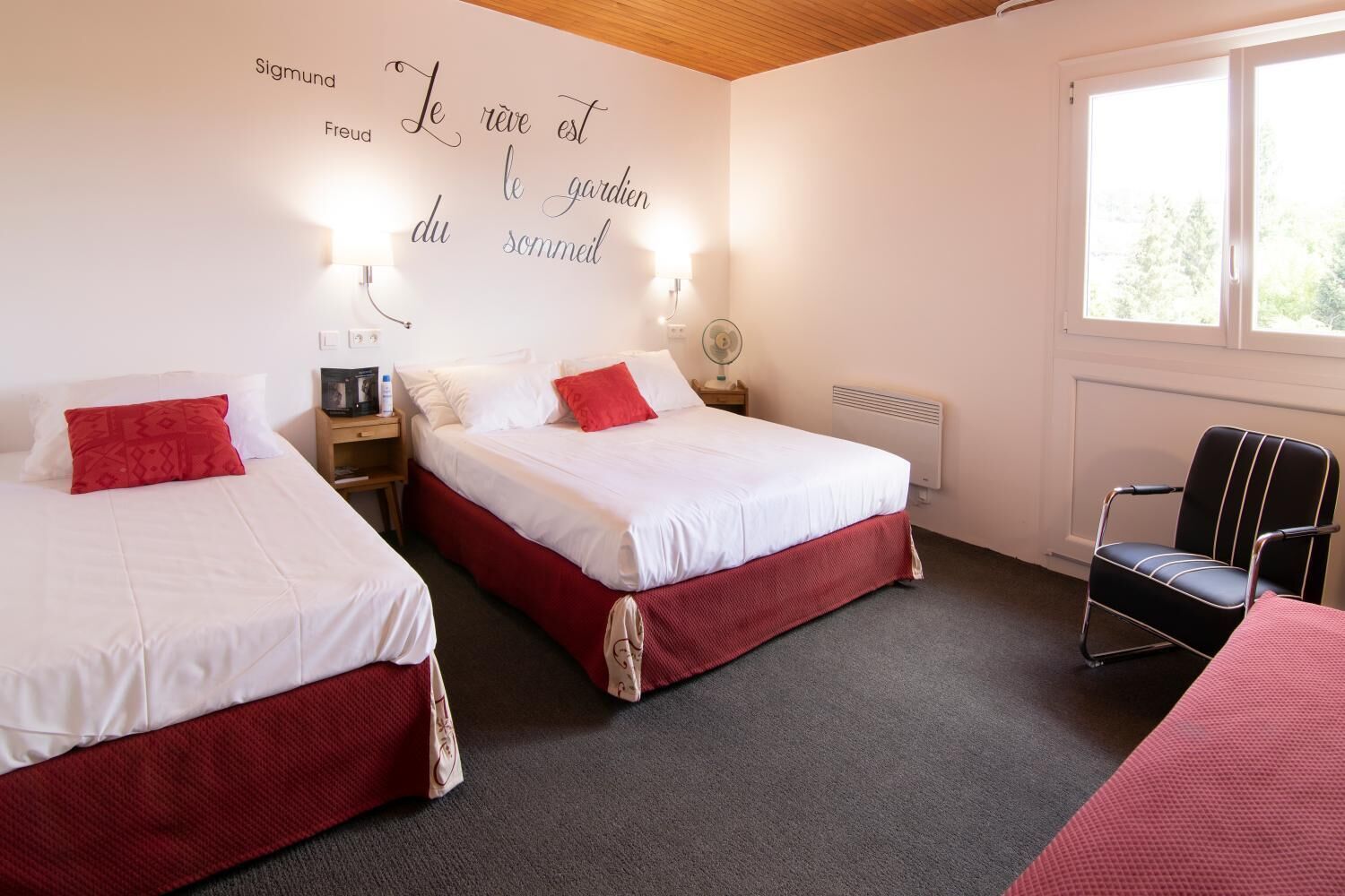 A hotel room with two beds, a chair, a wall clock, a heater, and a window.