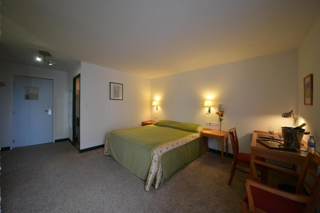 A hotel room with a bed, a desk, a chair, and a small table with a lamp.