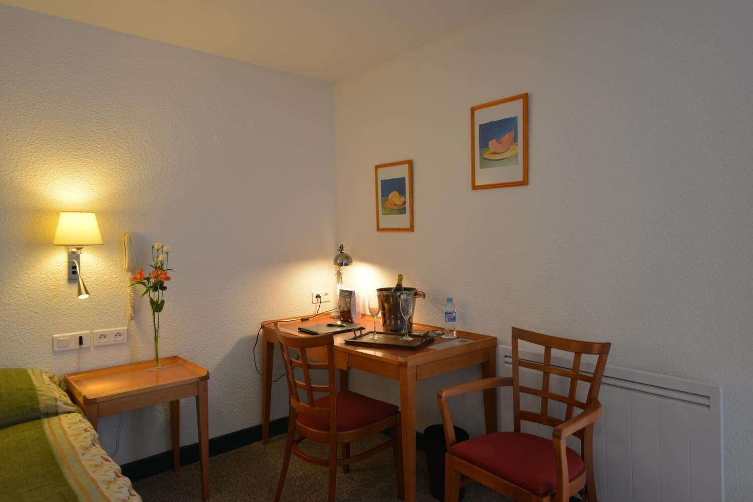 Een knus kamertje met een houten tafel en stoelen, een lamp, een vaas met bloemen en twee foto's in lijsten aan de muur.