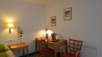 Een knus kamertje met een houten tafel en stoelen, een lamp, een vaas met bloemen en twee foto's in lijsten aan de muur.