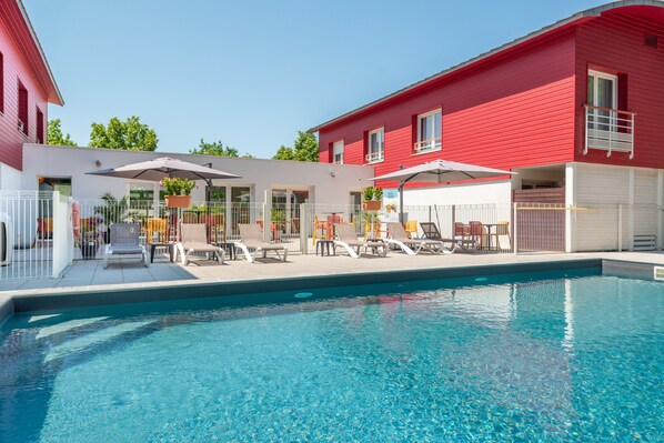 A swimming pool area with lounge chairs and umbrellas, a red building, and a covered seating area. - Comfort Aparthotel Nantes La Beaujoire (Nantes)