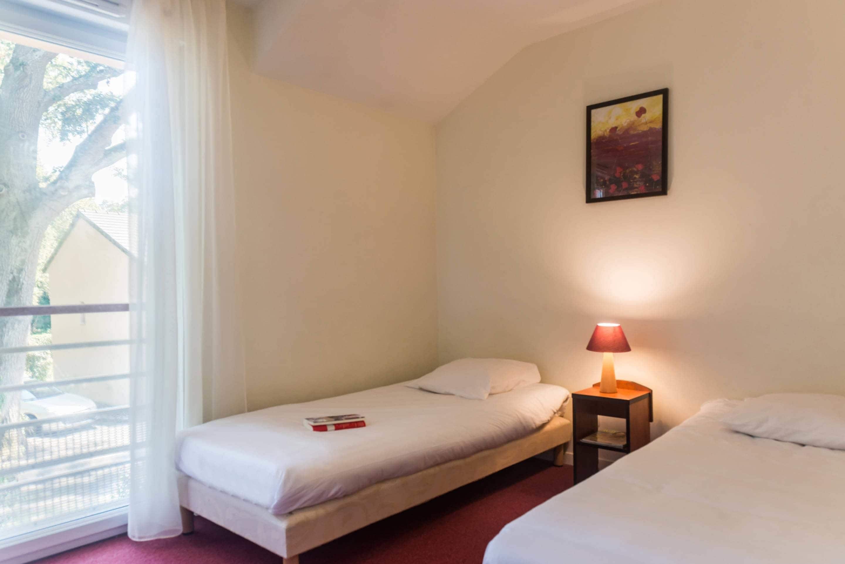 A hotel room with two beds, a small table with a lamp, a painting on the wall, and a window with curtains.