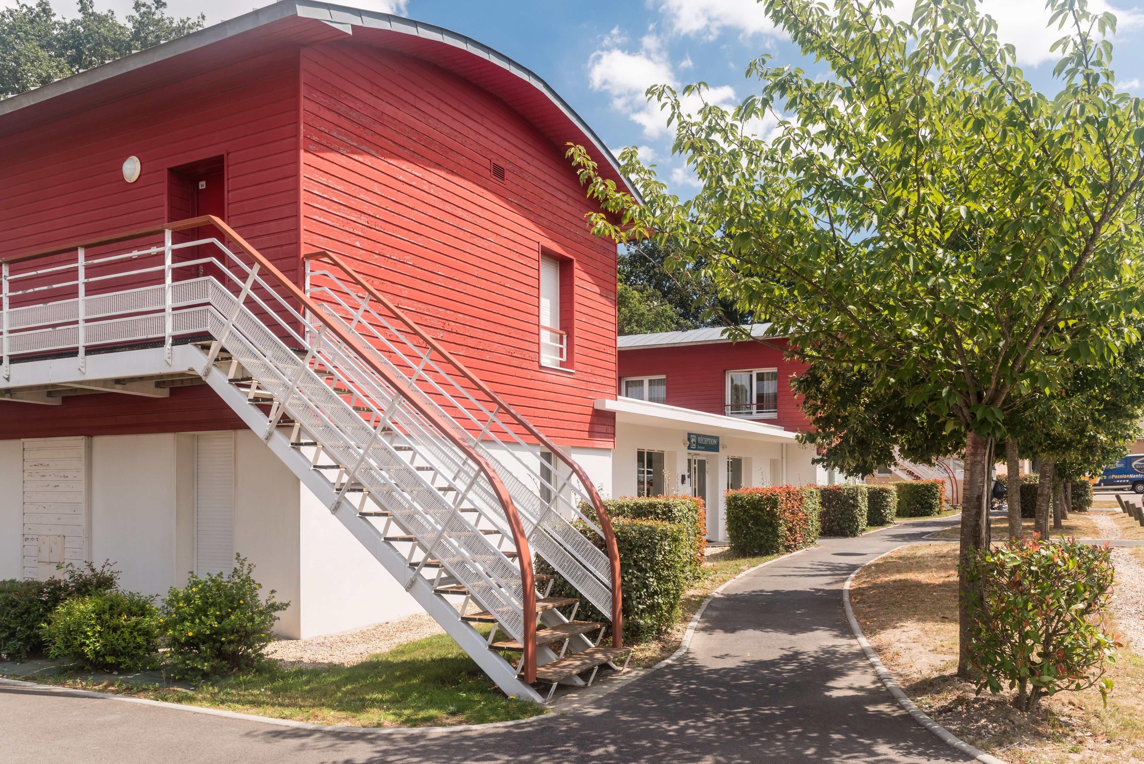 Un bâtiment rouge avec un escalier menant à une porte blanche, entouré de verdure et sous un ciel dégagé.