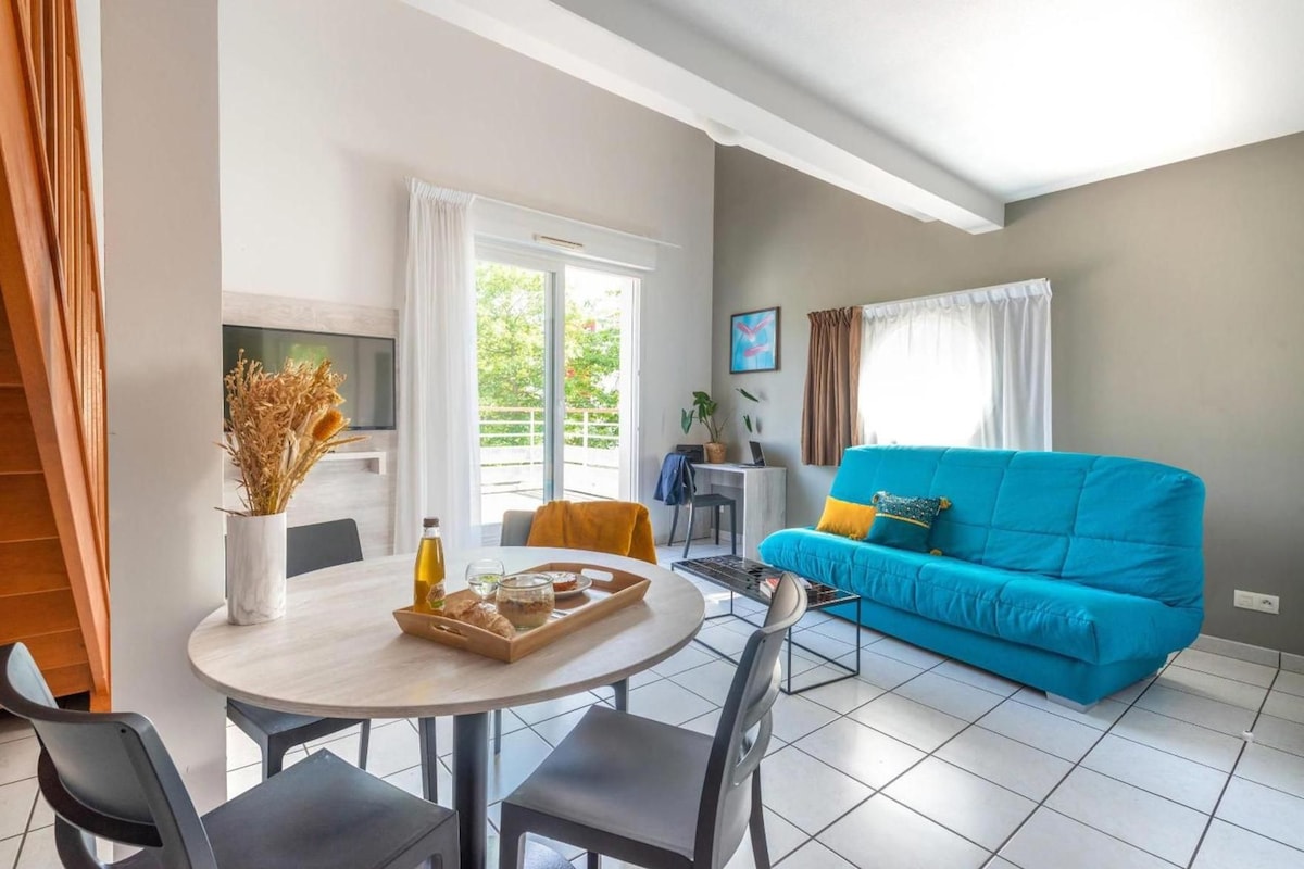 Una sala de estar moderna con un sofá azul, una mesa de comedor con sillas, un televisor montado en la pared y una ventana con cortinas.