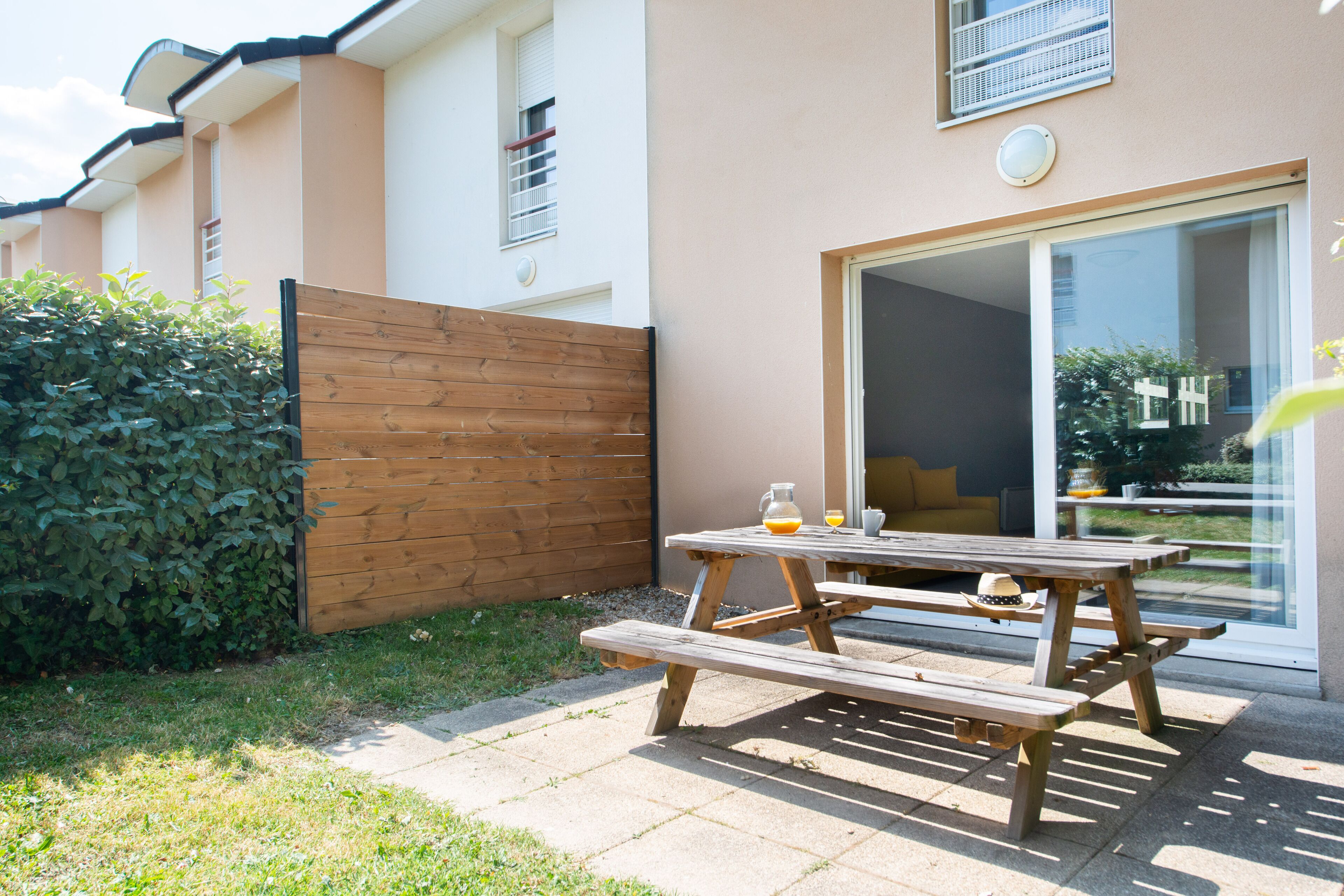 Een houten picknicktafel met bankjes in een achtertuin met een houten schutting en een schuifdeur die leidt naar een balkon.