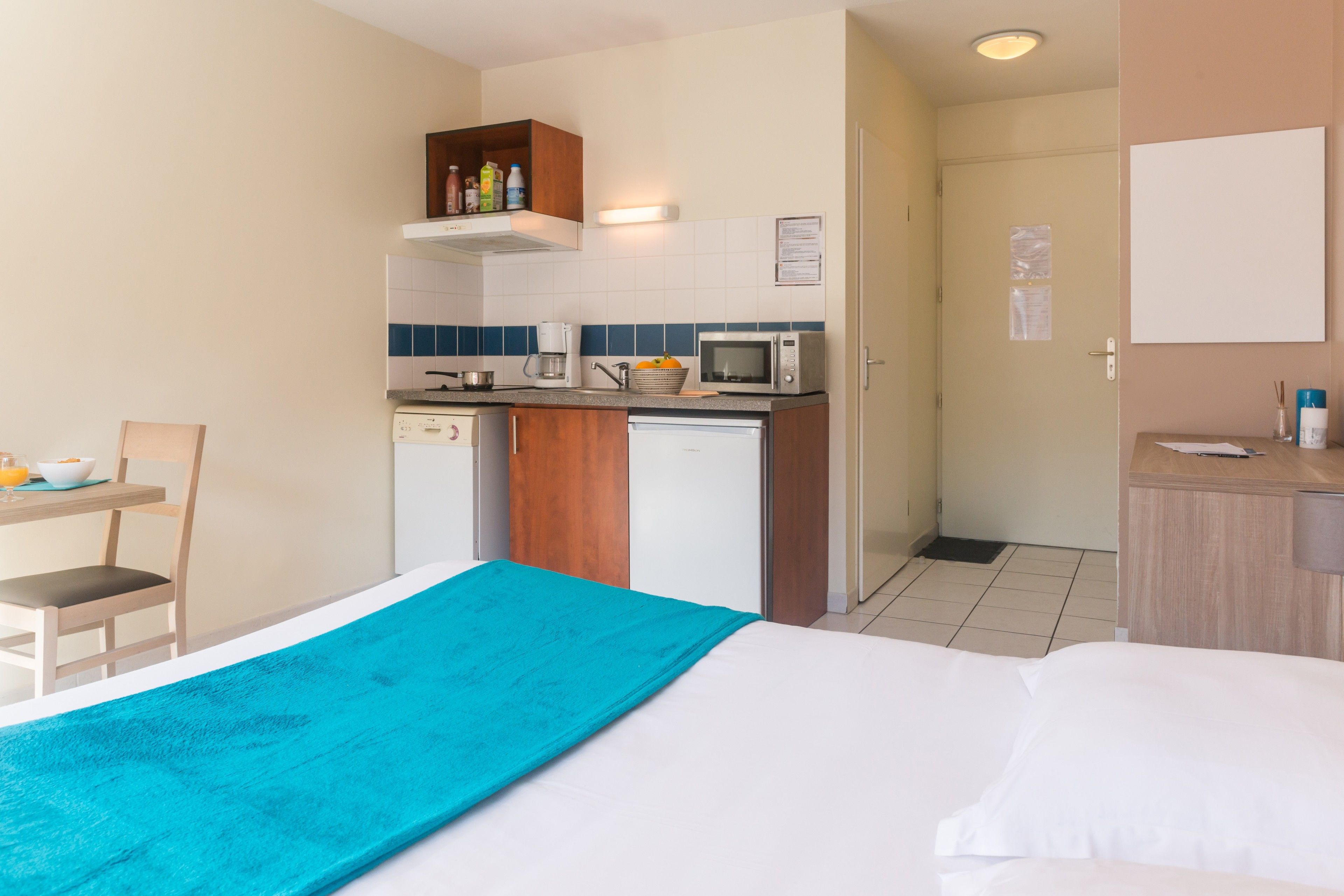 Een compacte hotelkamer met kitchenette, een bed met een blauwe deken, een kleine tafel met een stoel, en een deur naar een badkamer.