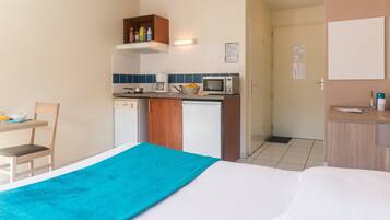 Een compacte hotelkamer met kitchenette, een bed met een blauwe deken, een kleine tafel met een stoel, en een deur naar een badkamer.