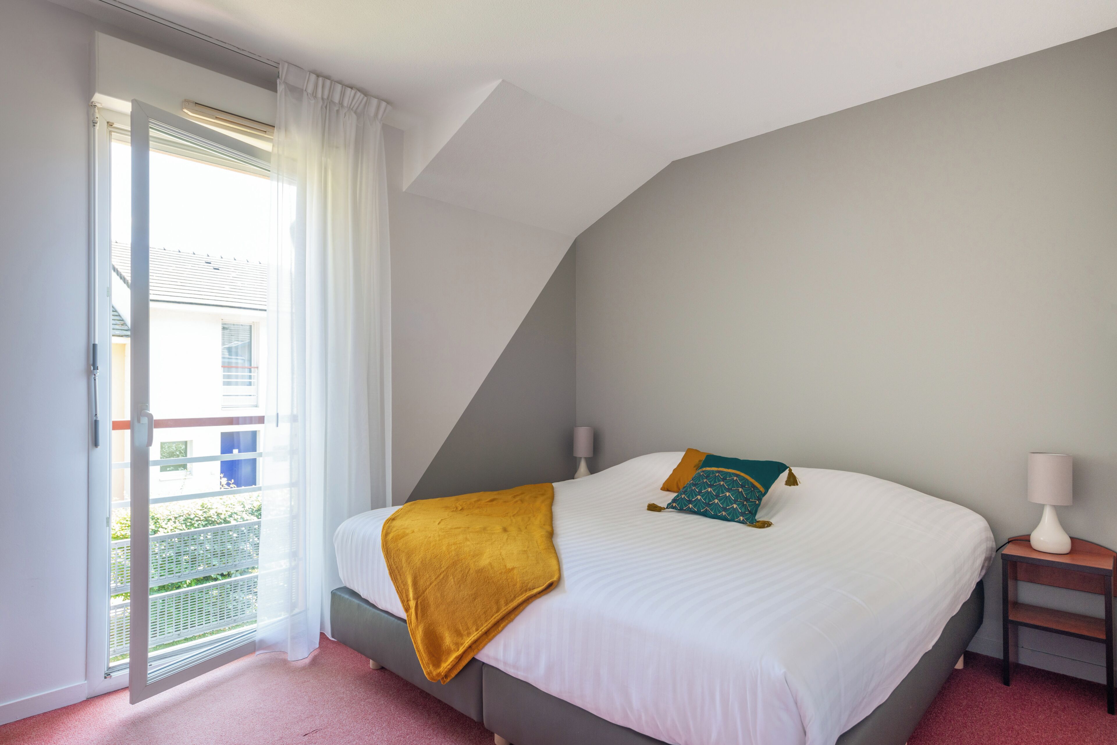 A bedroom with a bed, a nightstand, a window with curtains, and a view of the outside.