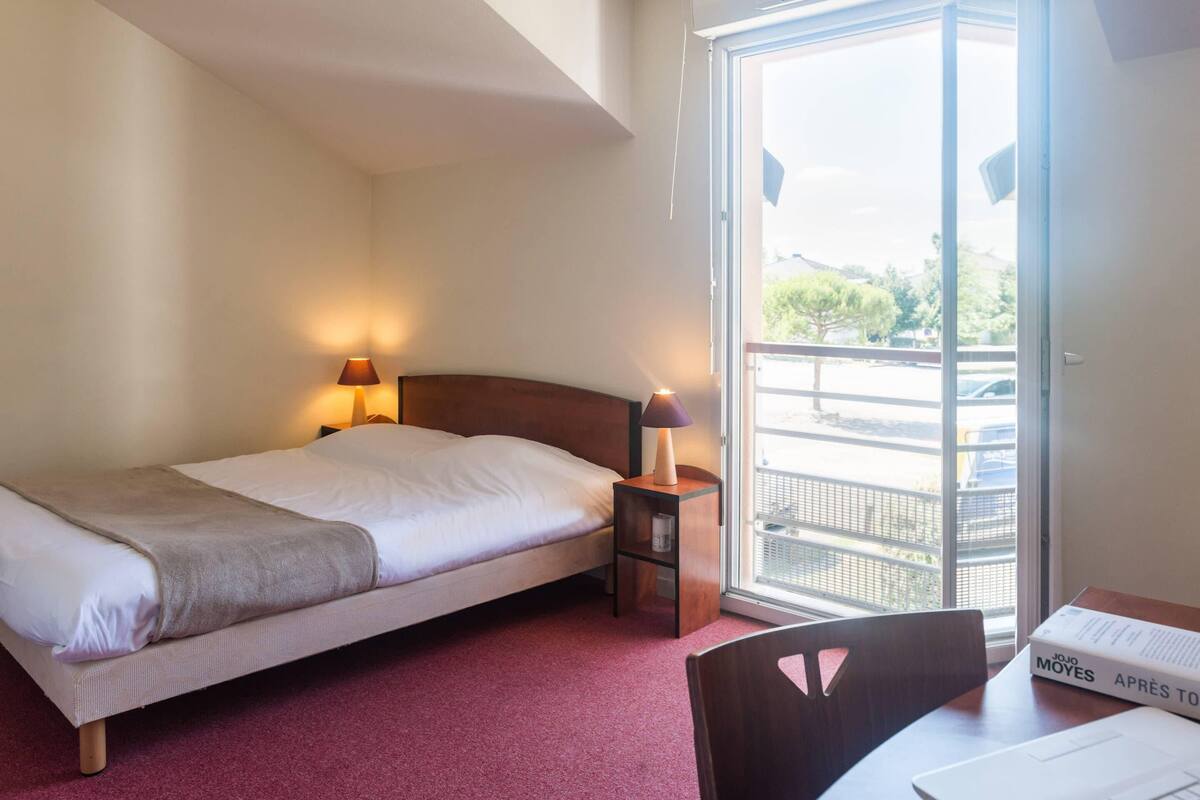 Une chambre d’hôtel avec un lit, des lampes de chevet, une petite table avec une chaise et une fenêtre donnant sur l’extérieur.