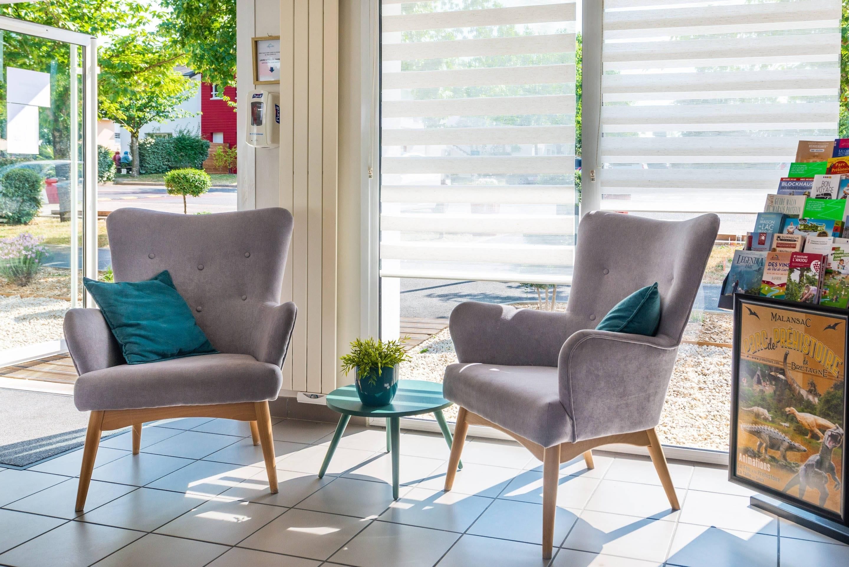 A modern interior with two grey armchairs, a small round table with a plant, and a window with blinds.