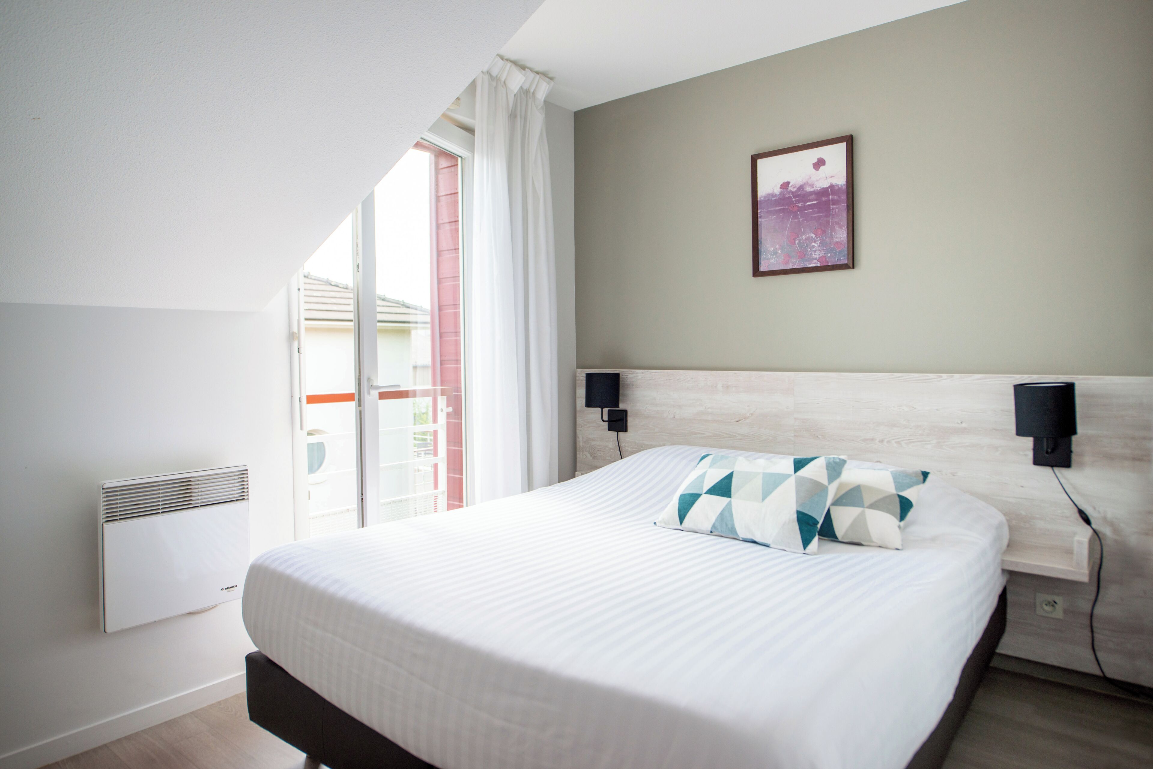 A bedroom with a bed, a wall-mounted TV, a picture, and a window with curtains.