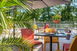Um pátio externo com mesa posta para o café da manhã, rodeado por cadeiras e vasos de plantas.