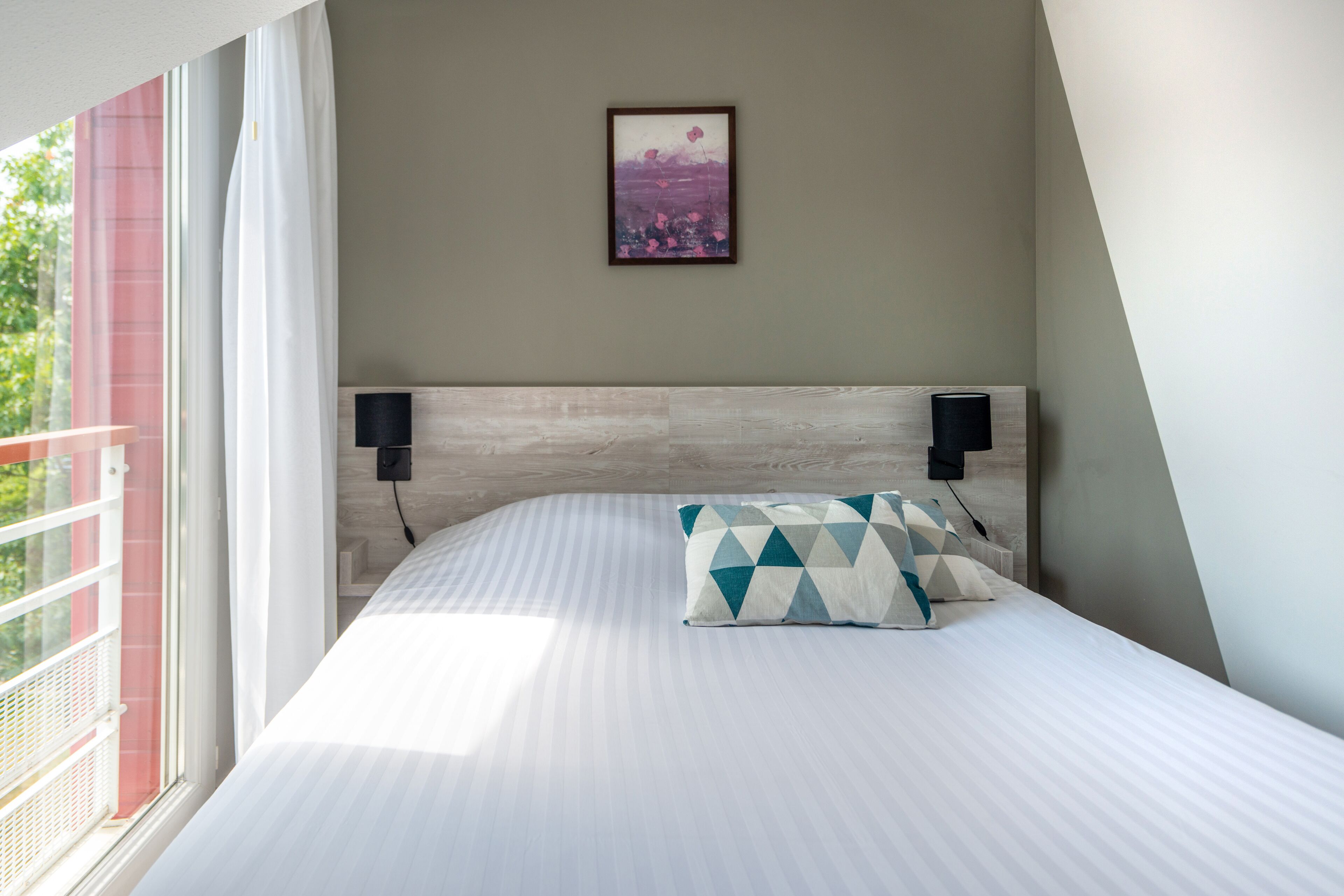 A bedroom with a large bed, a headboard with speakers, a framed picture on the wall, and a balcony with a railing.