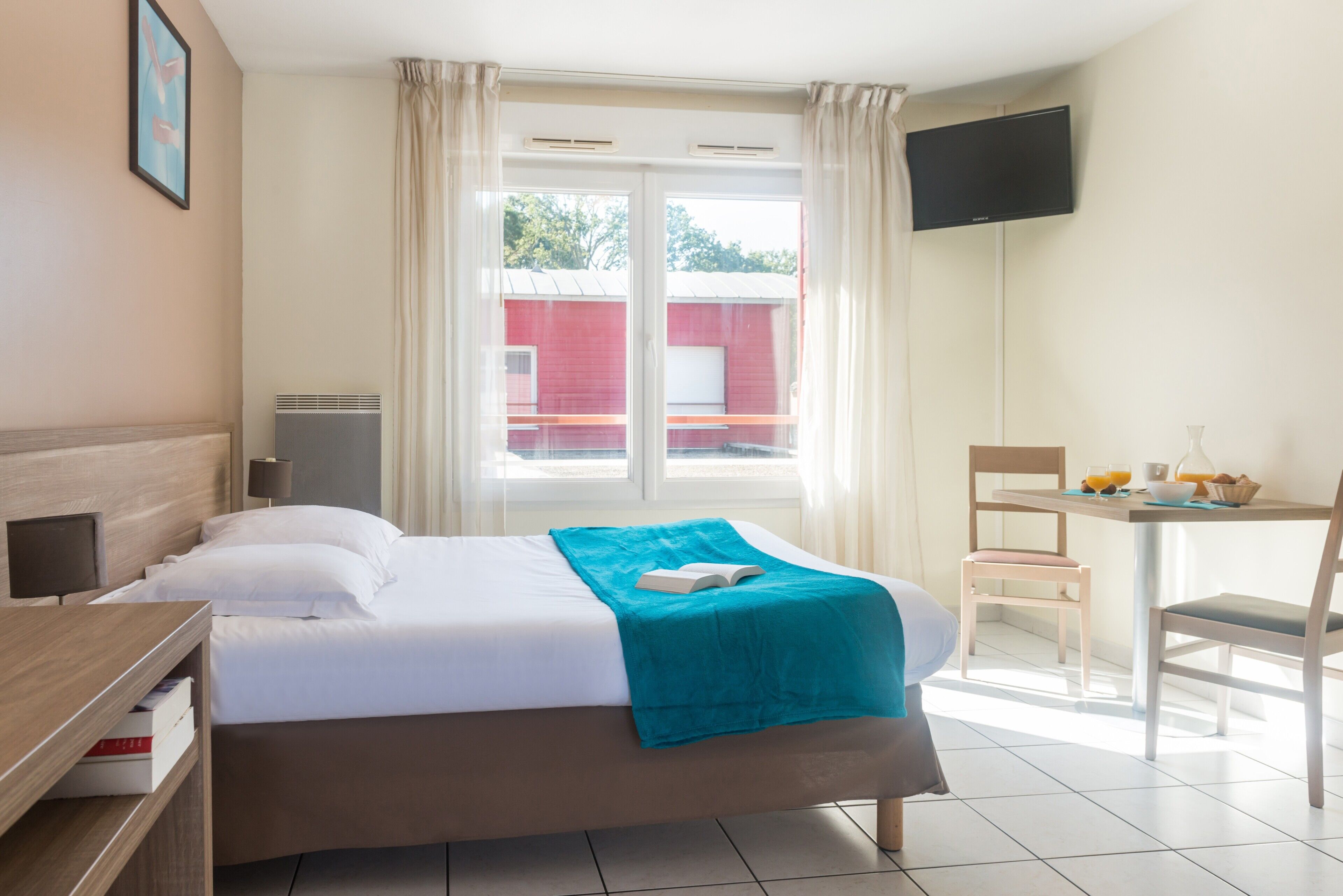 A hotel room with a bed, a desk with a chair, a television, and a window with curtains.