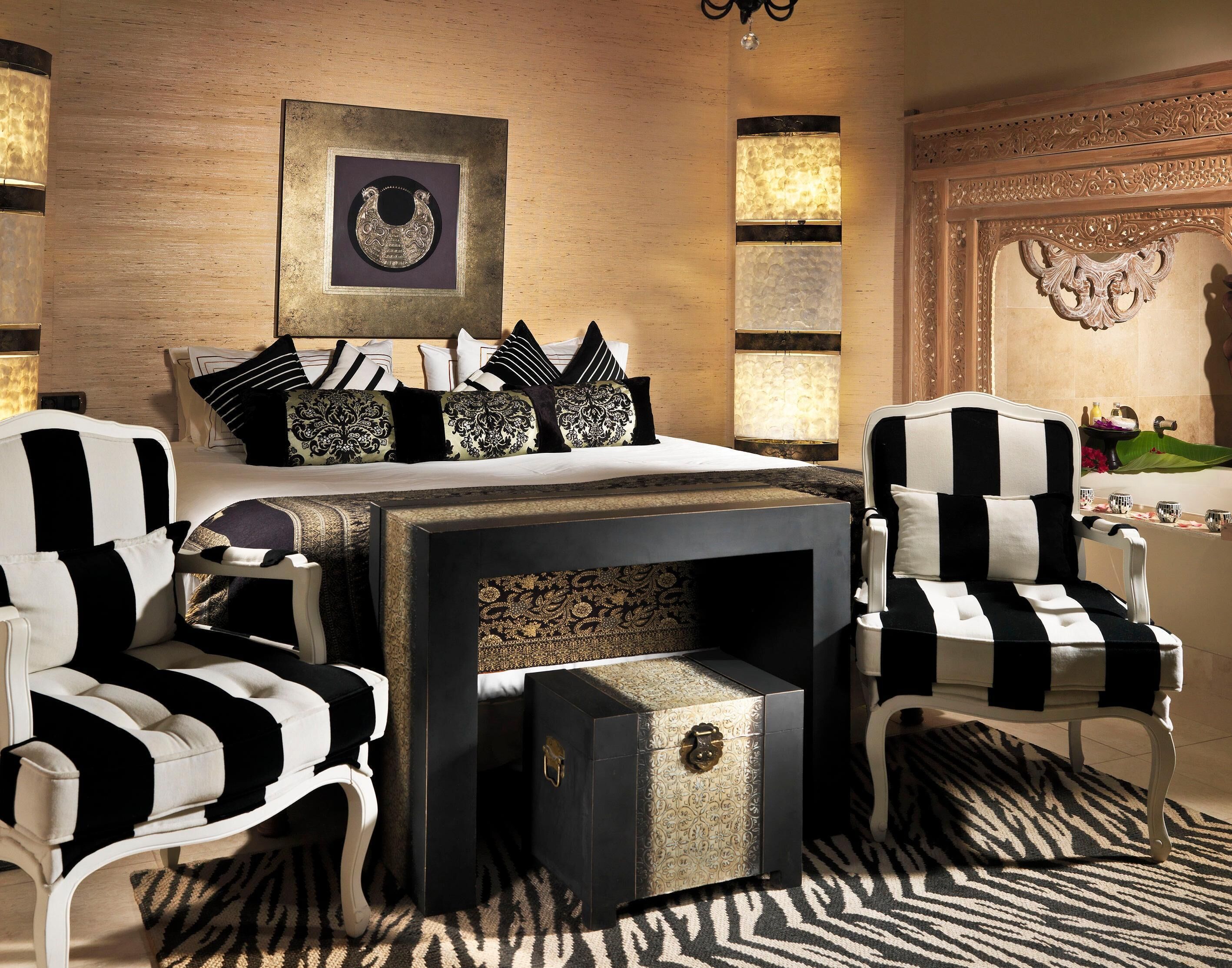 A bedroom with a bed, two armchairs, a nightstand, and a decorative wall piece.