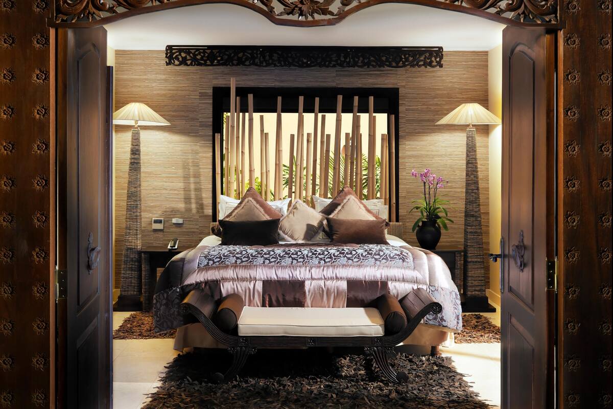 A bedroom with a bed, pillows, a bedside table, a lamp, and a potted plant.