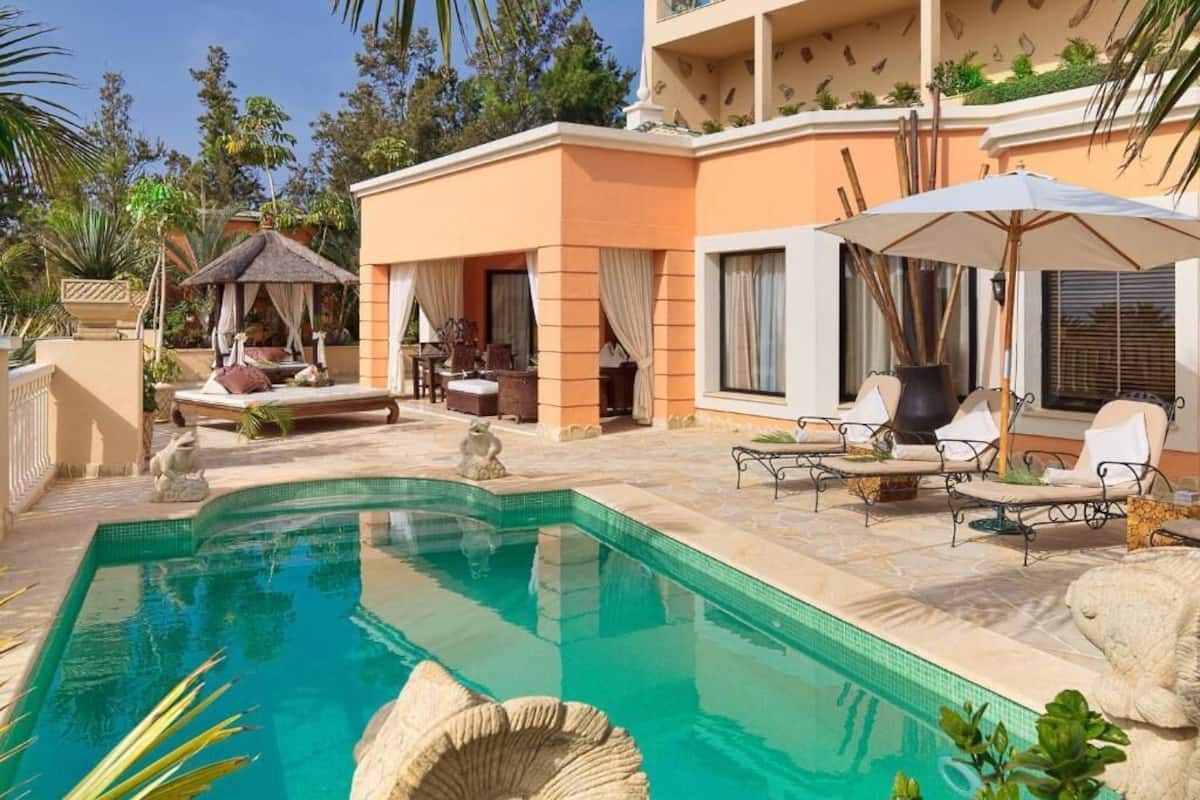 A pool area with lounge chairs and umbrellas, a thatched-roof gazebo, and a stone sculpture.