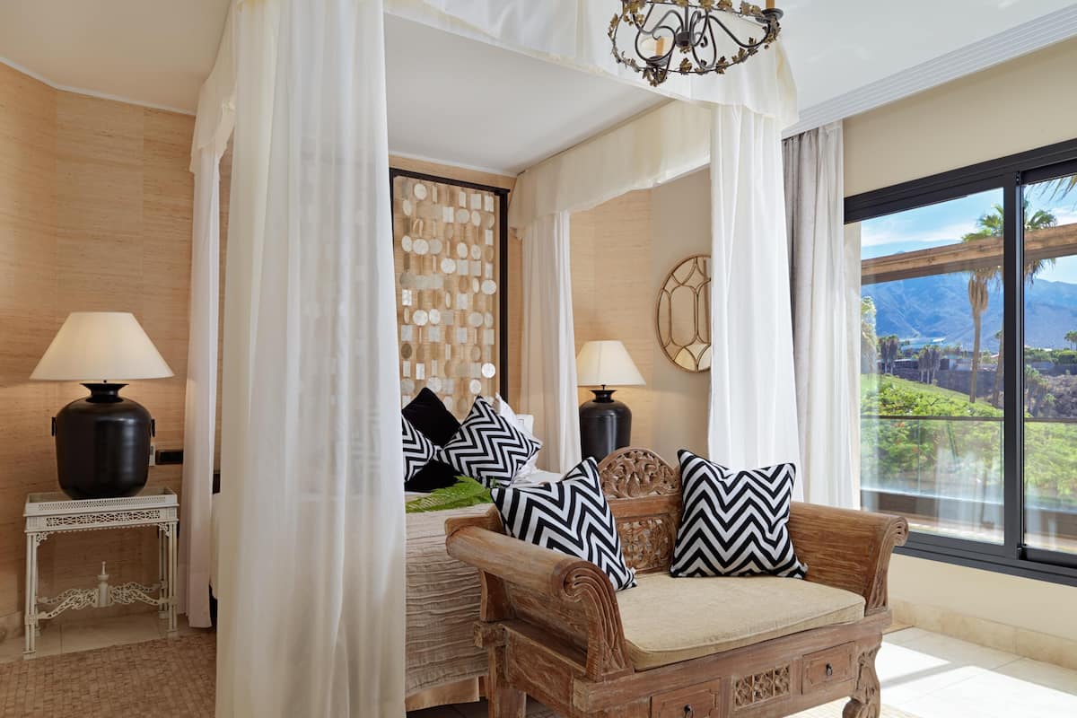 A bedroom with a bed, a wooden bench with cushions, a decorative wall, and a large window with a view.