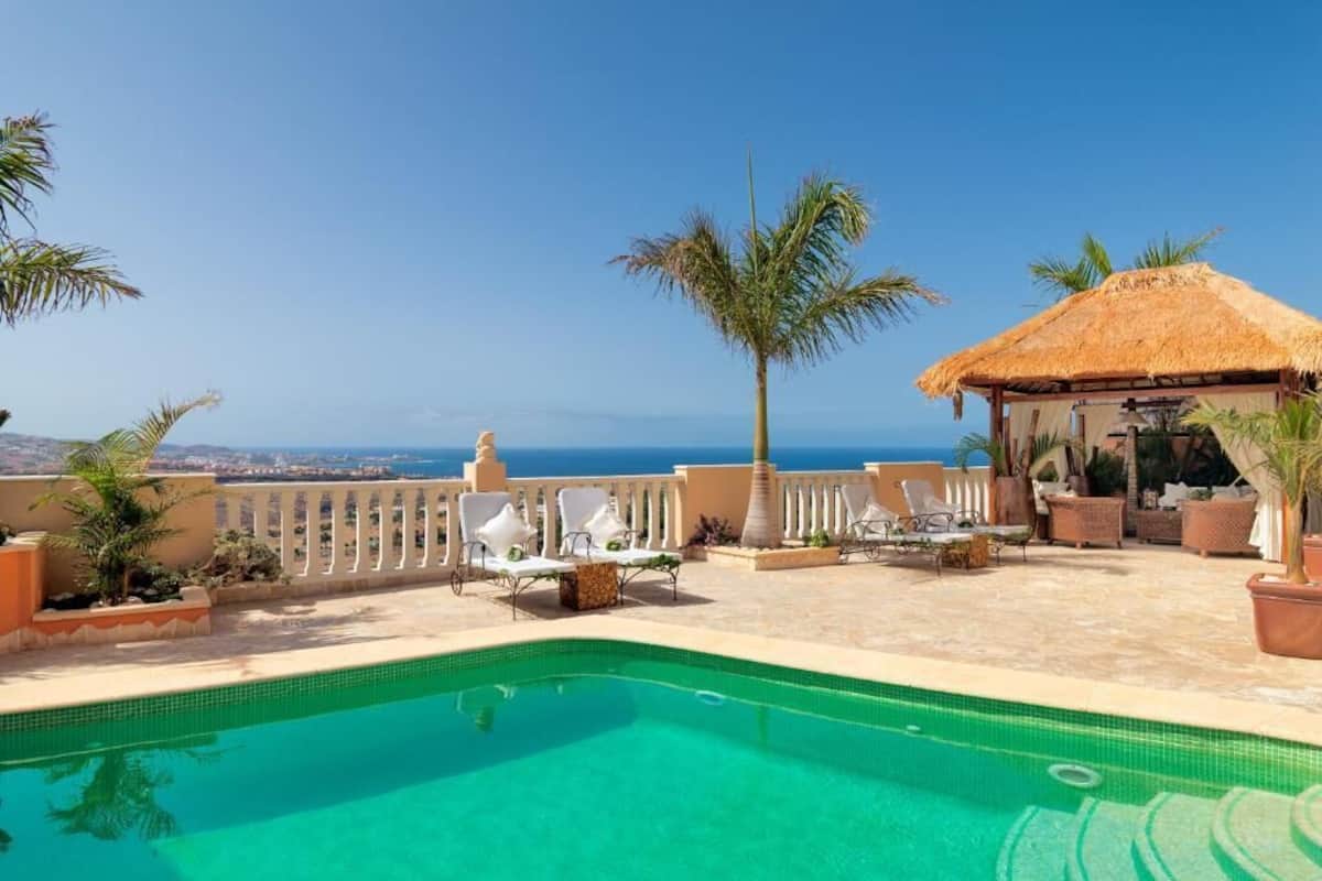 A swimming pool with lounge chairs, a thatched-roof structure, and a view of the ocean.