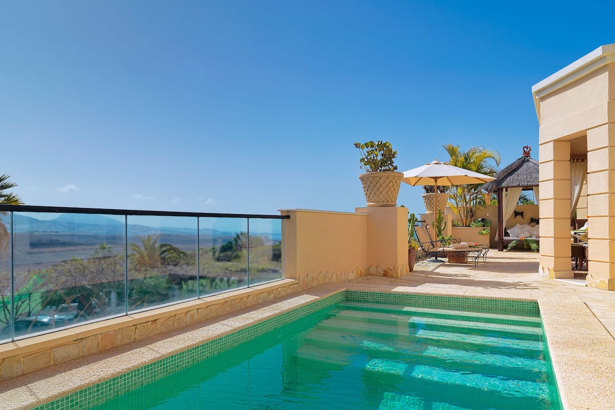 A pool with a glass barrier, a lounge area with a thatched roof, and a view of distant hills.