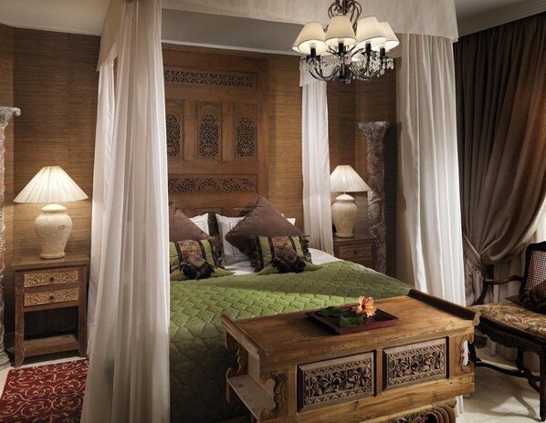 Un dormitorio con cabecera de madera, colcha acolchada verde, varias almohadas, una mesita de noche con lámpara y una mesa con una flor.