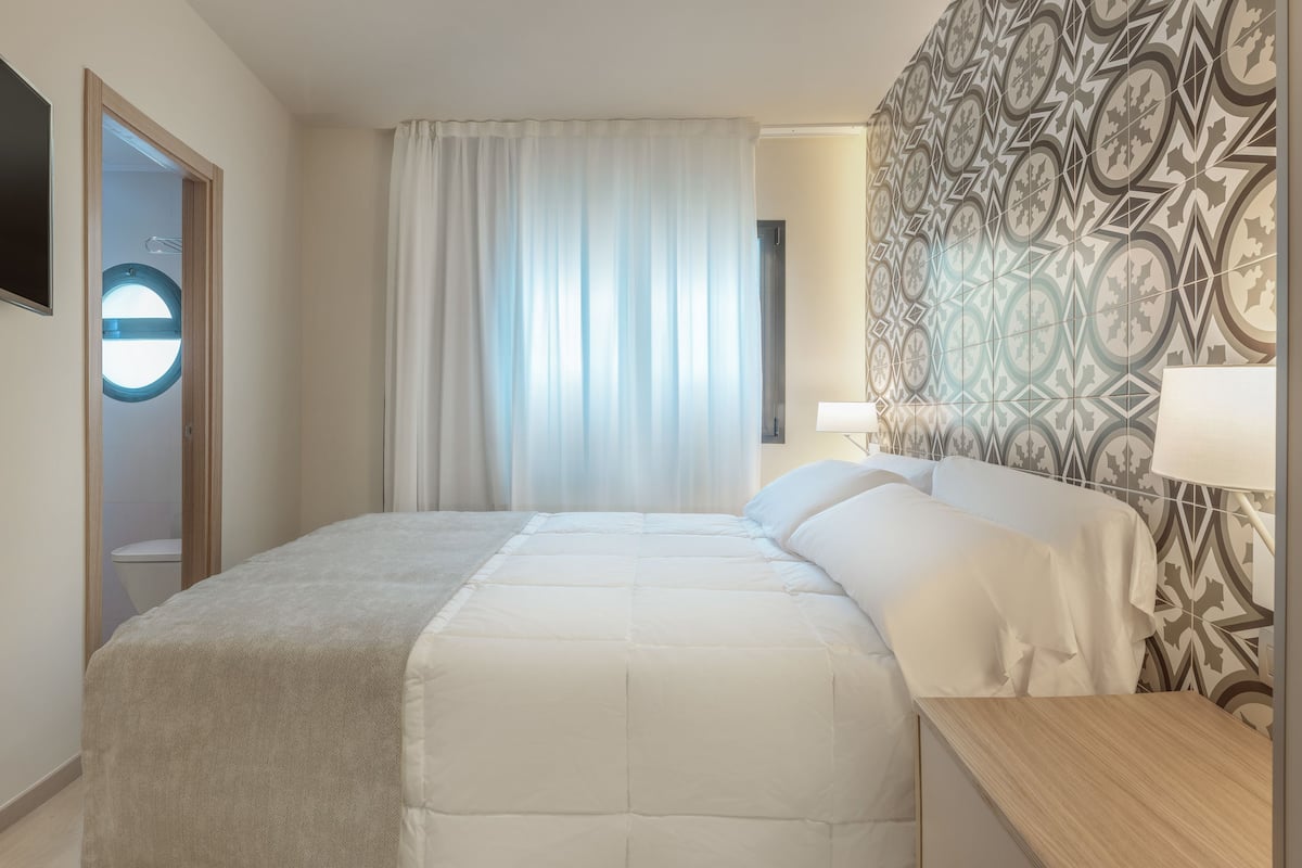 A hotel room with a bed, a bedside table, a lamp, a window with curtains, and a patterned wall.