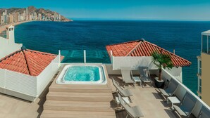 Un área de piscina en la azotea con un jacuzzi, sillones de descanso y vistas al océano y al horizonte de la ciudad.