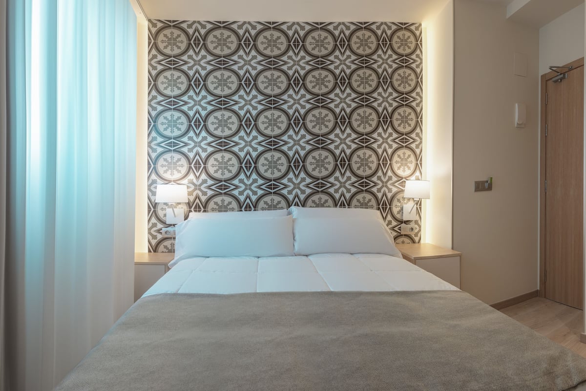 A hotel room with a bed, bedside lamps, a patterned wallpaper, a wardrobe, and a small table with a lamp.