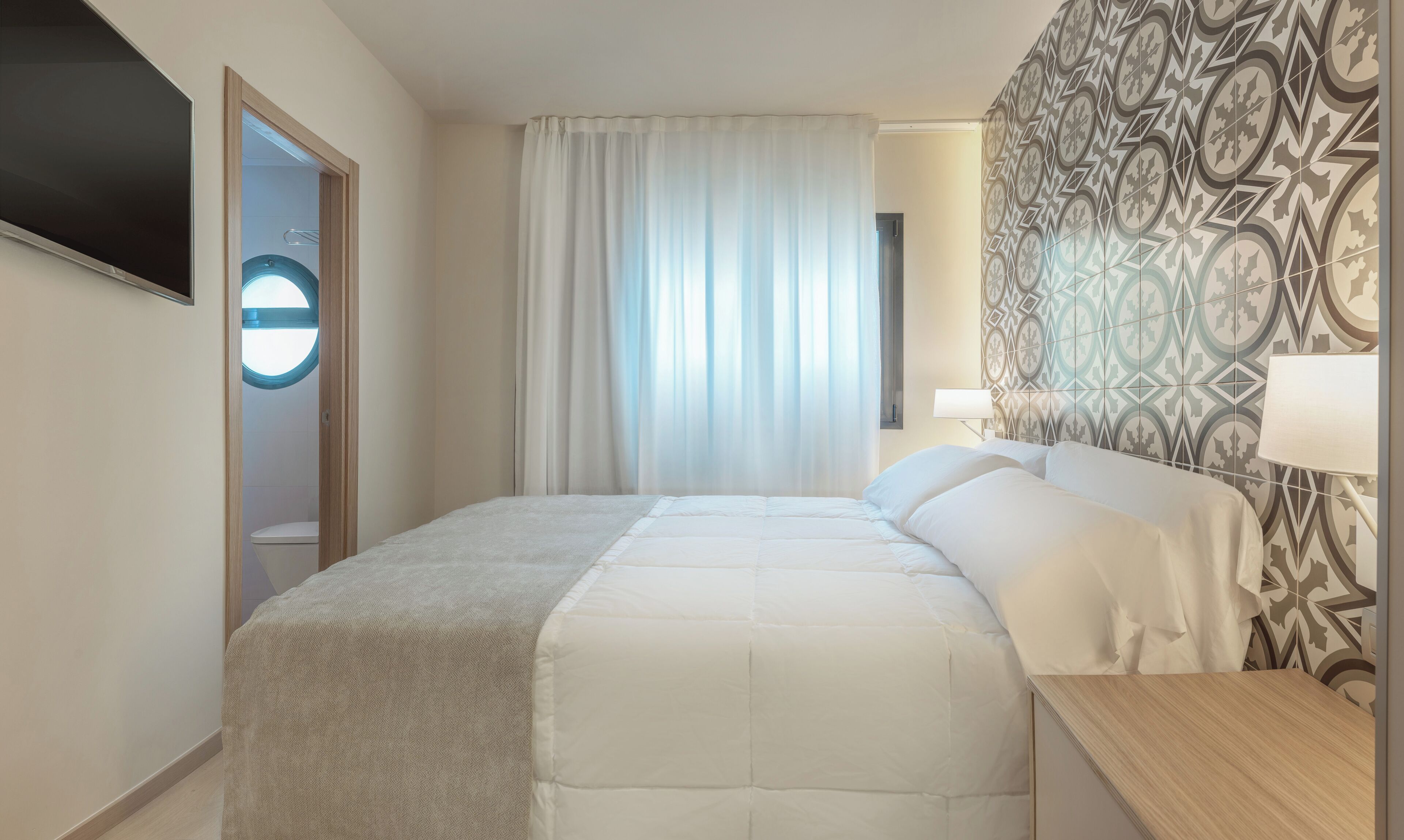A hotel room with a bed, a bedside table, a lamp, a window with curtains, and a patterned wall.