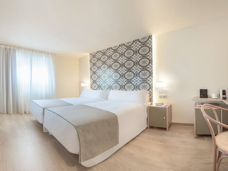 A hotel room with a bed, bedside tables, a desk, a chair, a TV, and a patterned wall.