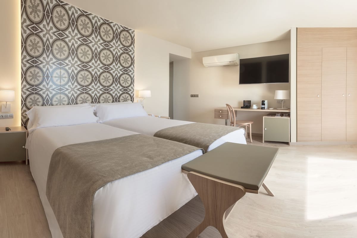 A modern hotel room with two beds, a small desk, a TV, and a balcony with a city view.
