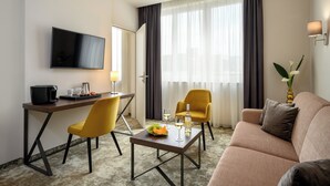 A hotel room with a flat-screen TV, a desk with a coffee maker, two yellow chairs, a brown sofa, and a dining table with a bowl of fruit and wine glasses. - Leonardo Boutique Hotel Budapest M-Square (Budapest)
