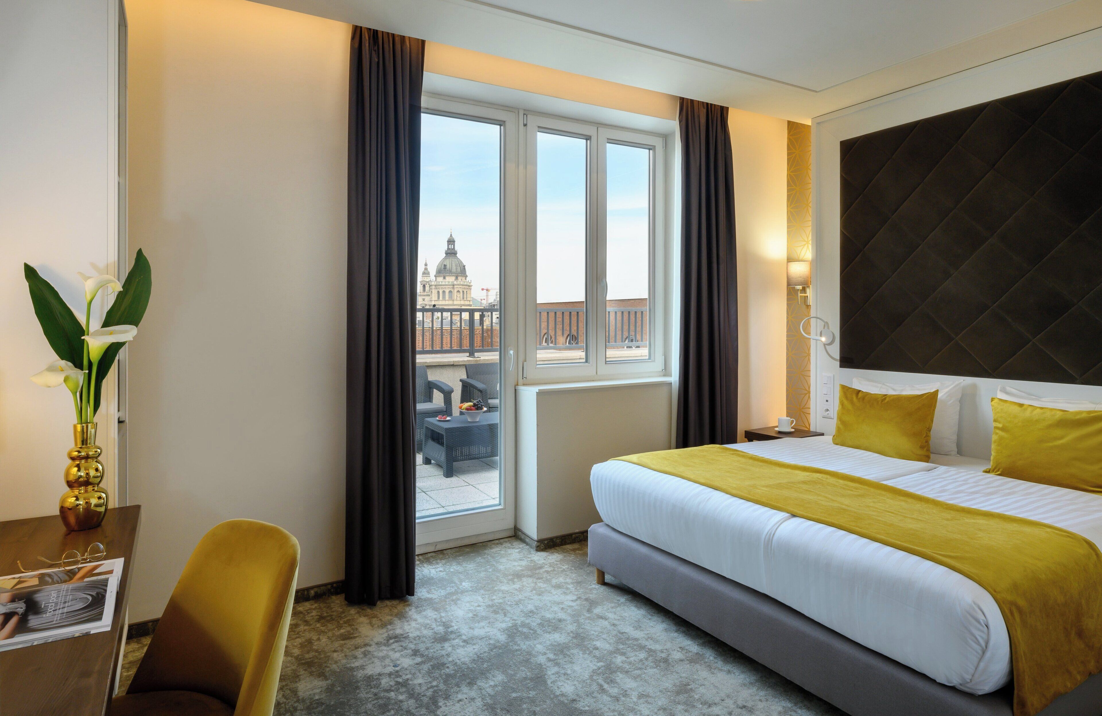 A hotel room with a large bed, a desk with a chair, a vase of flowers, and a view of a building through the window.