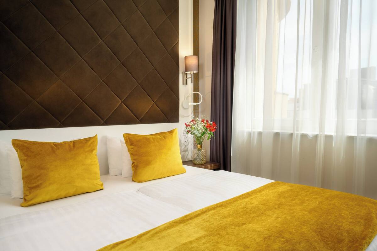 A hotel room with a large bed, two yellow pillows, a white bedspread, a brown headboard, a vase with flowers, and sheer curtains.