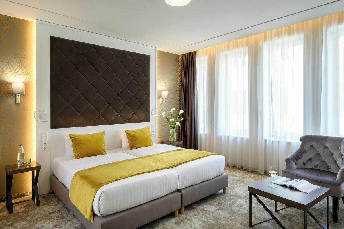 A modern hotel room with a large bed, a grey armchair, a small table, and a large window with curtains.