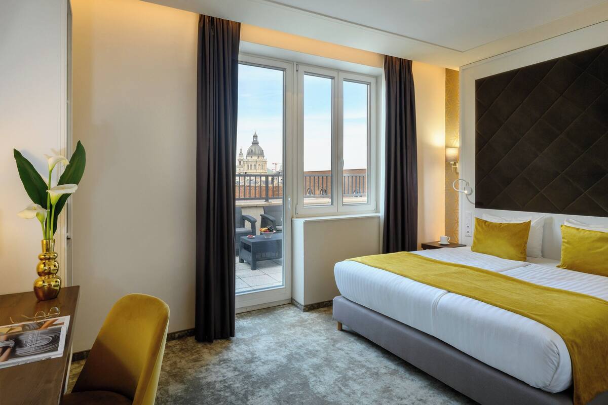 A hotel room with a large bed, a desk with a chair, a vase of flowers, and a view of a building through the window.
