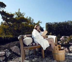 Una persona asseguda en un banc de fusta a l'aire lliure, amb un llibre, envoltada de roques i vegetació.