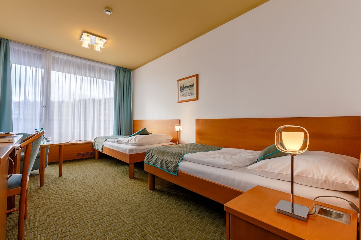 Quarto de hotel com duas camas de solteiro, uma escrivaninha, uma cadeira, uma mesa de cabeceira com abajur e uma janela com cortinas.