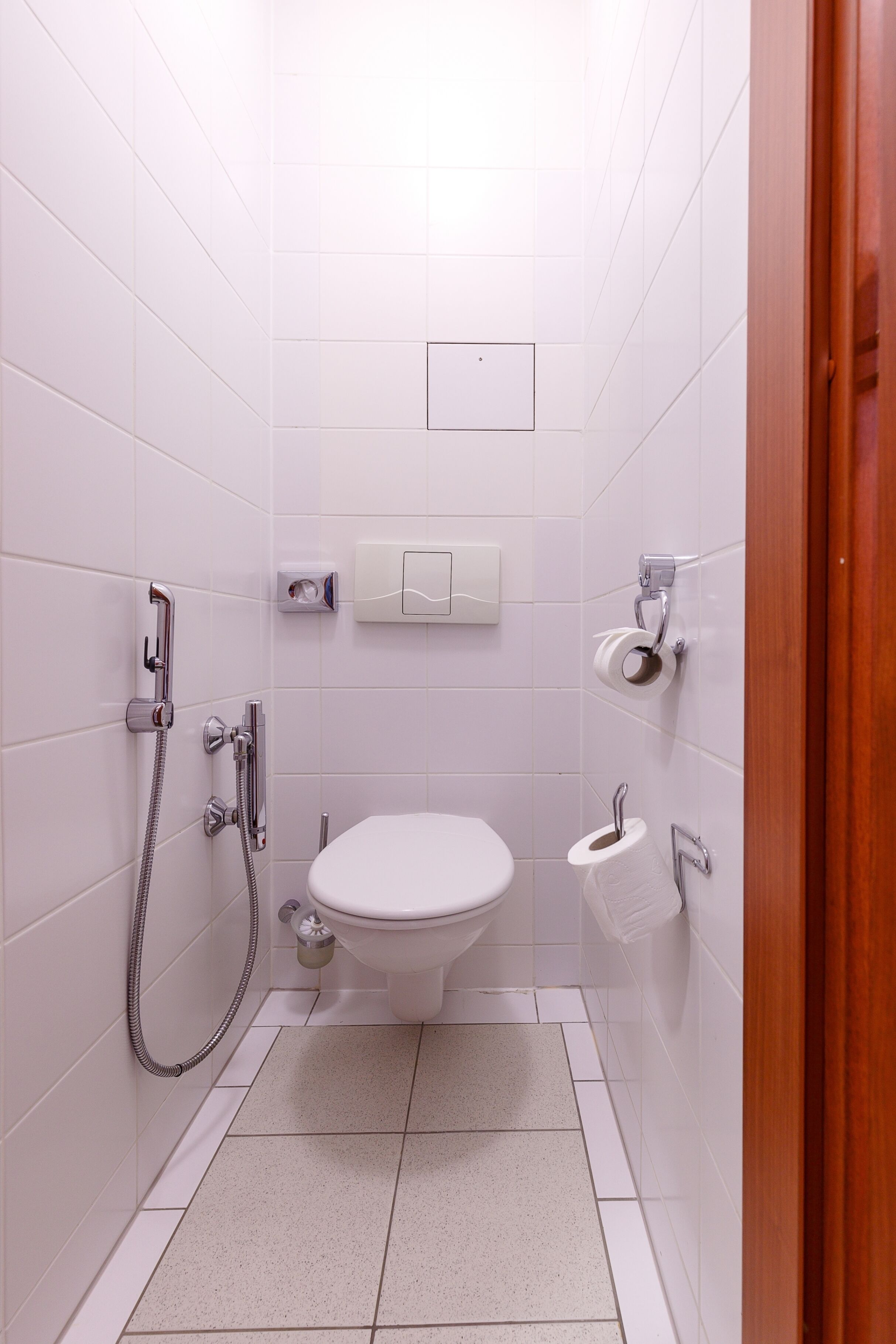 A small, clean bathroom with white tiles, a toilet, a shower, and a towel dispenser.