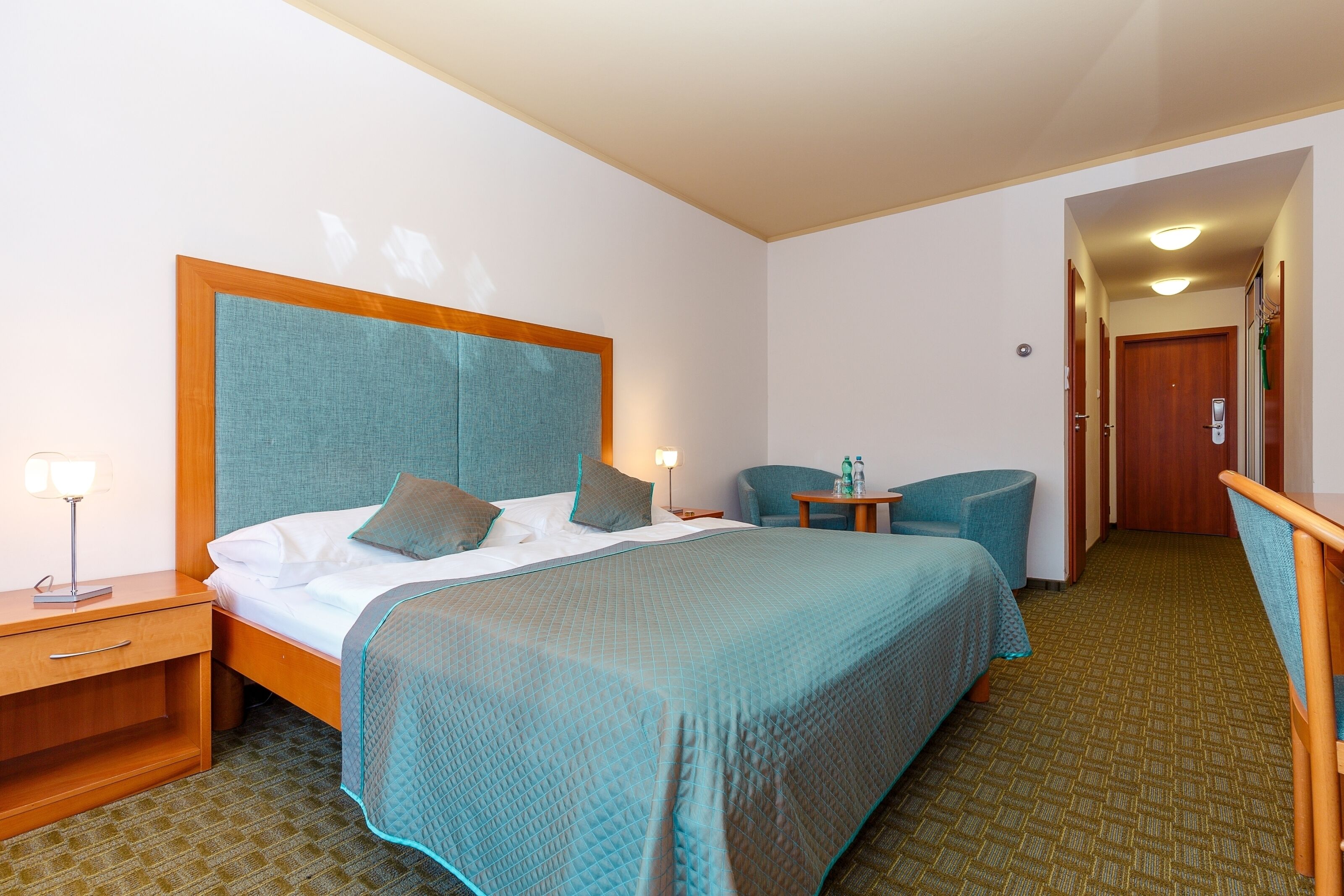 Habitación de hotel con una cama grande, una mesita de noche con lámpara, una pequeña mesa con una botella y una silla.