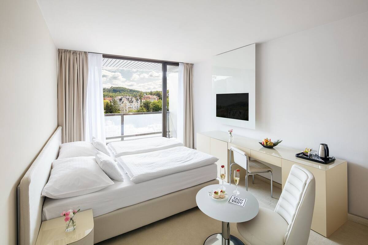 Quarto de hotel com cama, escrivaninha, cadeira, mesinha com um copo de vinho e televisão na parede.