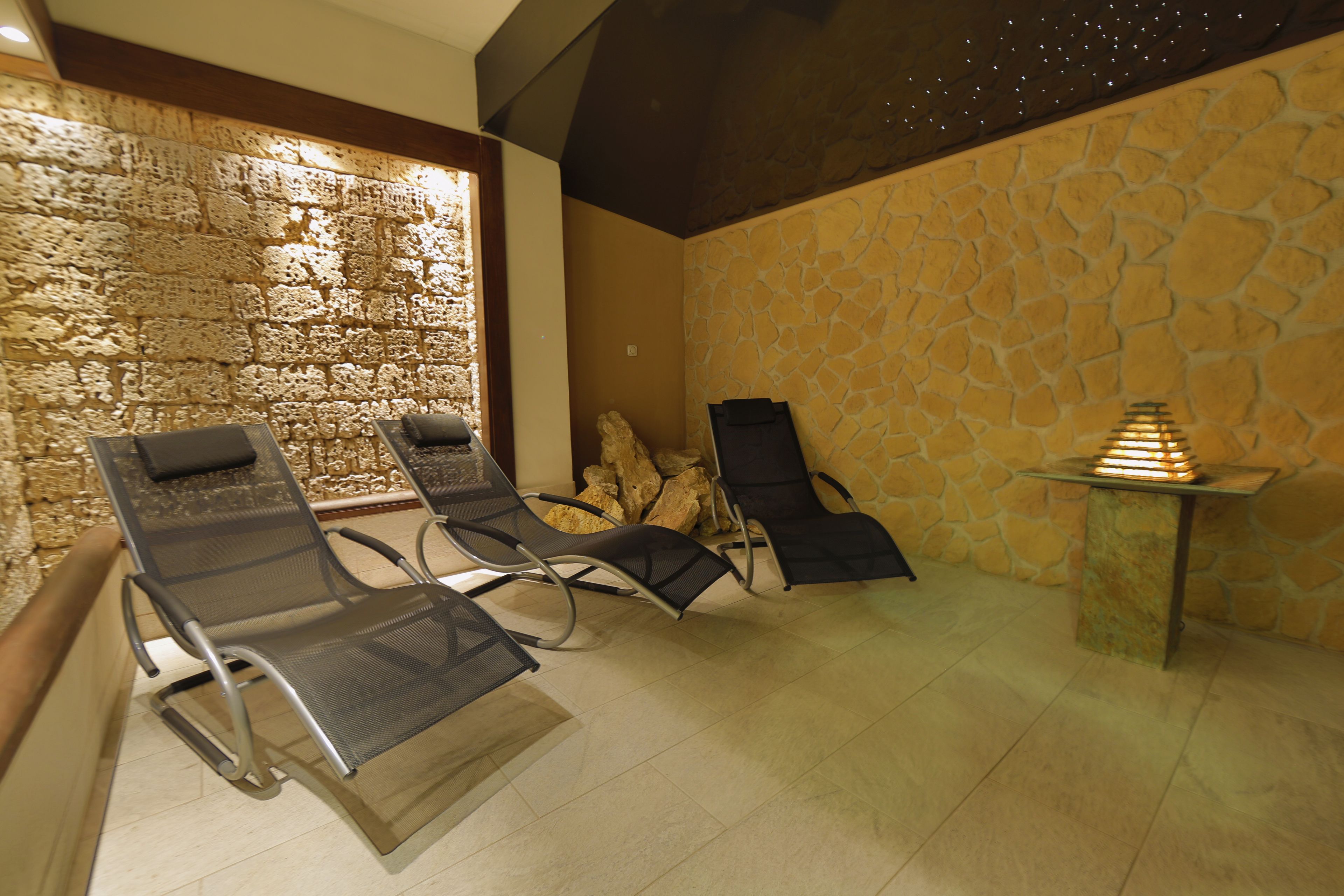 A spa area with stone walls, two massage chairs, a small table with a lamp, and a tiled floor.